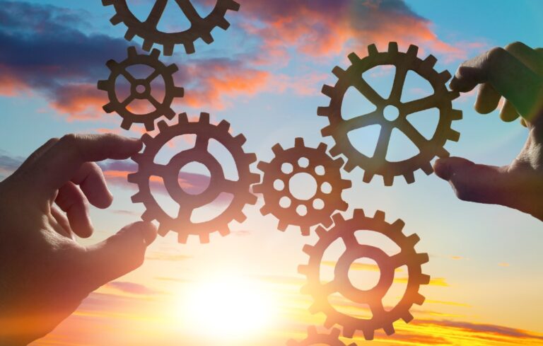 Hands collect gear in a puzzle against the sky in the sunset.