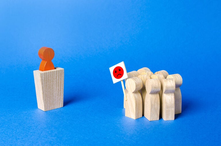 Crowd of wooden figures with posters expresses distrust for other wooden figure on a podium